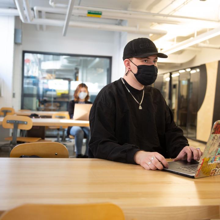 A masked student sit at a desk, looking at their laptop screen