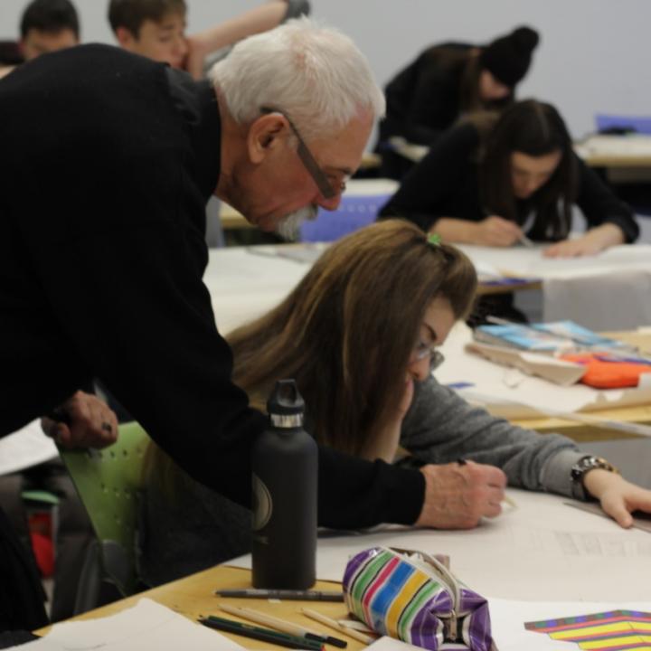 A teacher leans over to assist student with their drawing. 